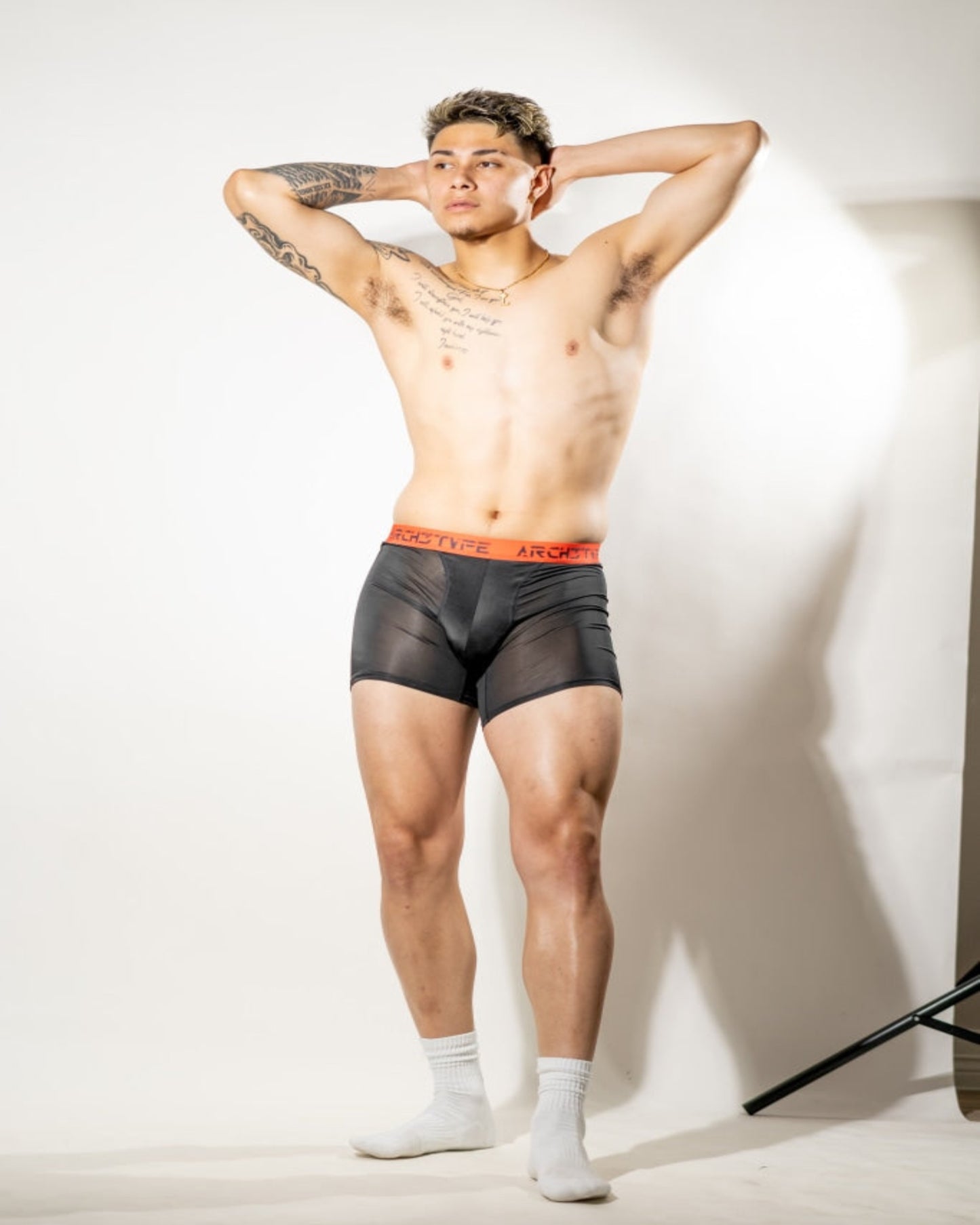 A shirtless male model with tattoos poses in black mesh boxer briefs with a red waistband and white socks against a white studio backdrop.