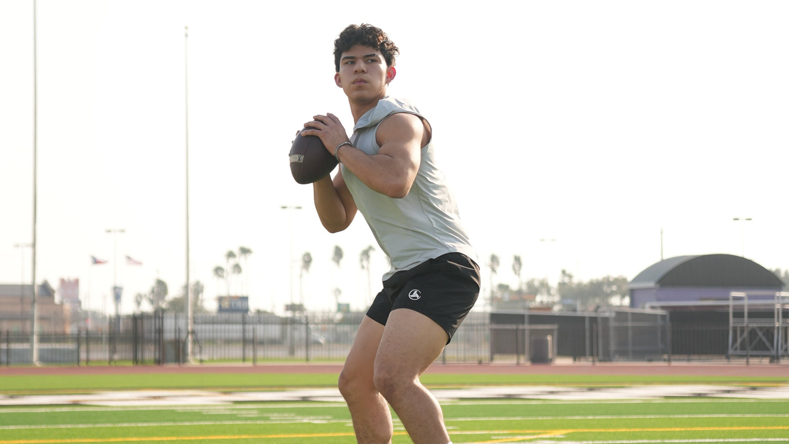 A man wearing a light gray sleeveless shirt, black athletic shorts, white socks, and white sneakers is running on a green football field while holding a football.